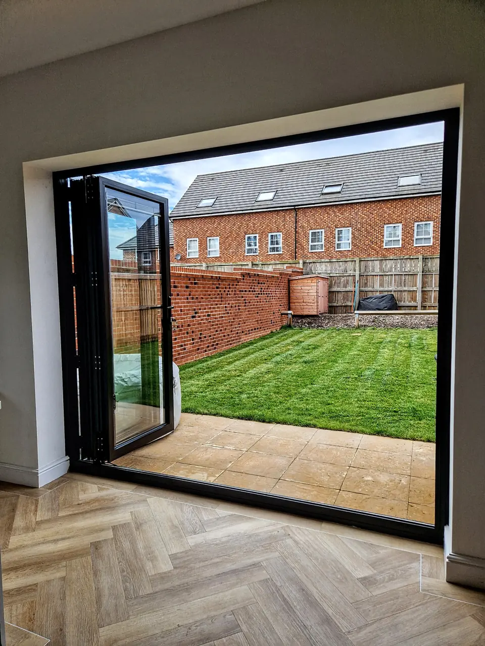 The same three-leaf black aluminium bifold from one fixed position inside an unfurnished room with a herringbone floor: closed with the integral blinds raised, part-folded, then folded fully back and stacked flat against the left reveal, leaving the opening onto the lawn completely clear.